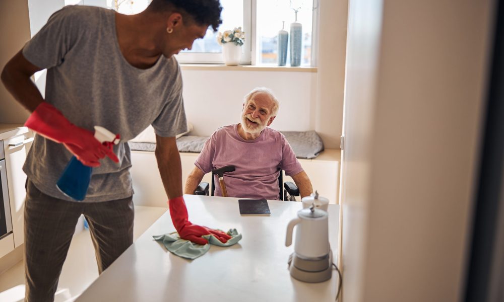 Smiling pleased disabled aged Caucasian person looking at the volunteer dusting the table in his presence